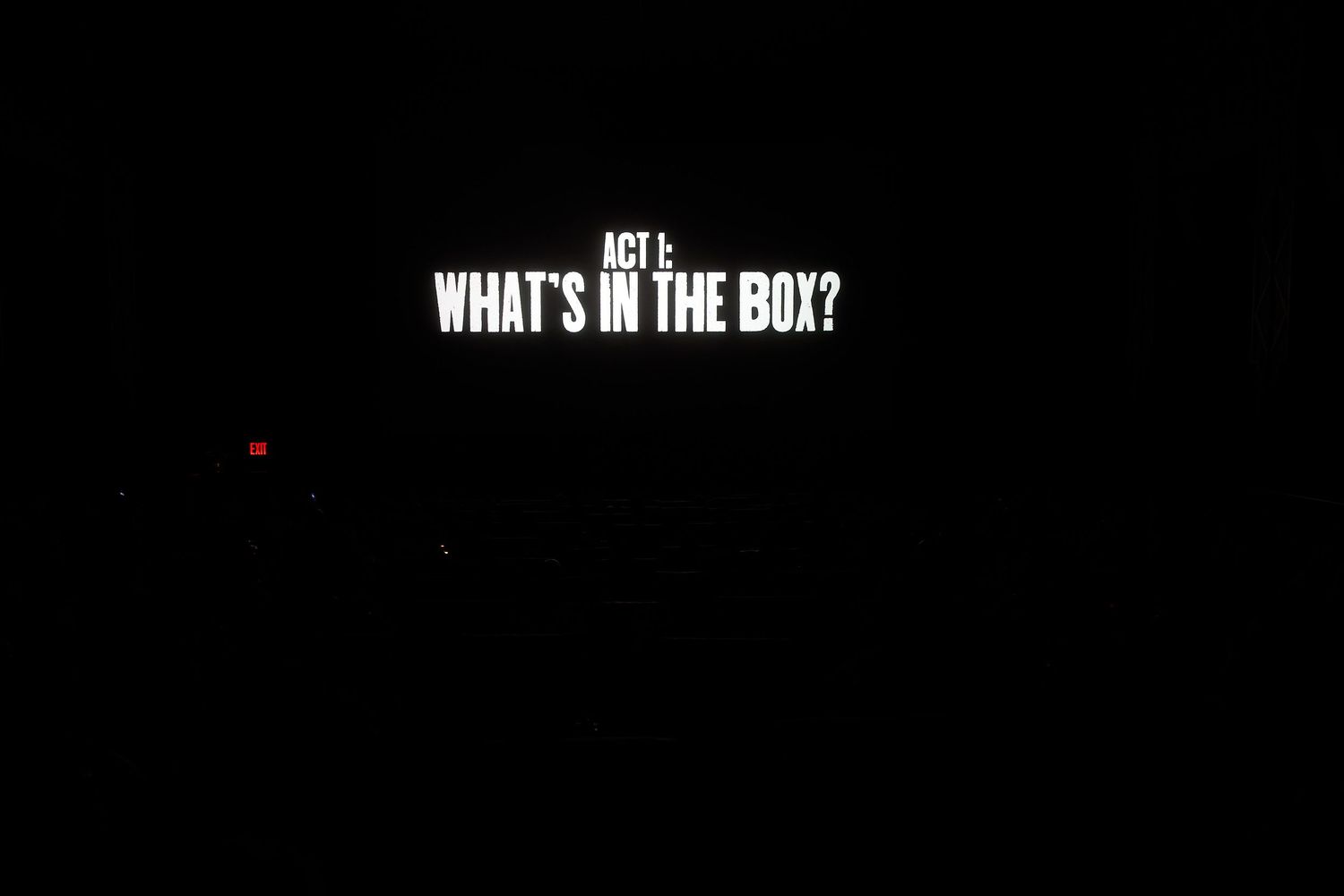 Dark screen displaying bold white text reading Act 1: What's In The Box? during a theatrical presentation.