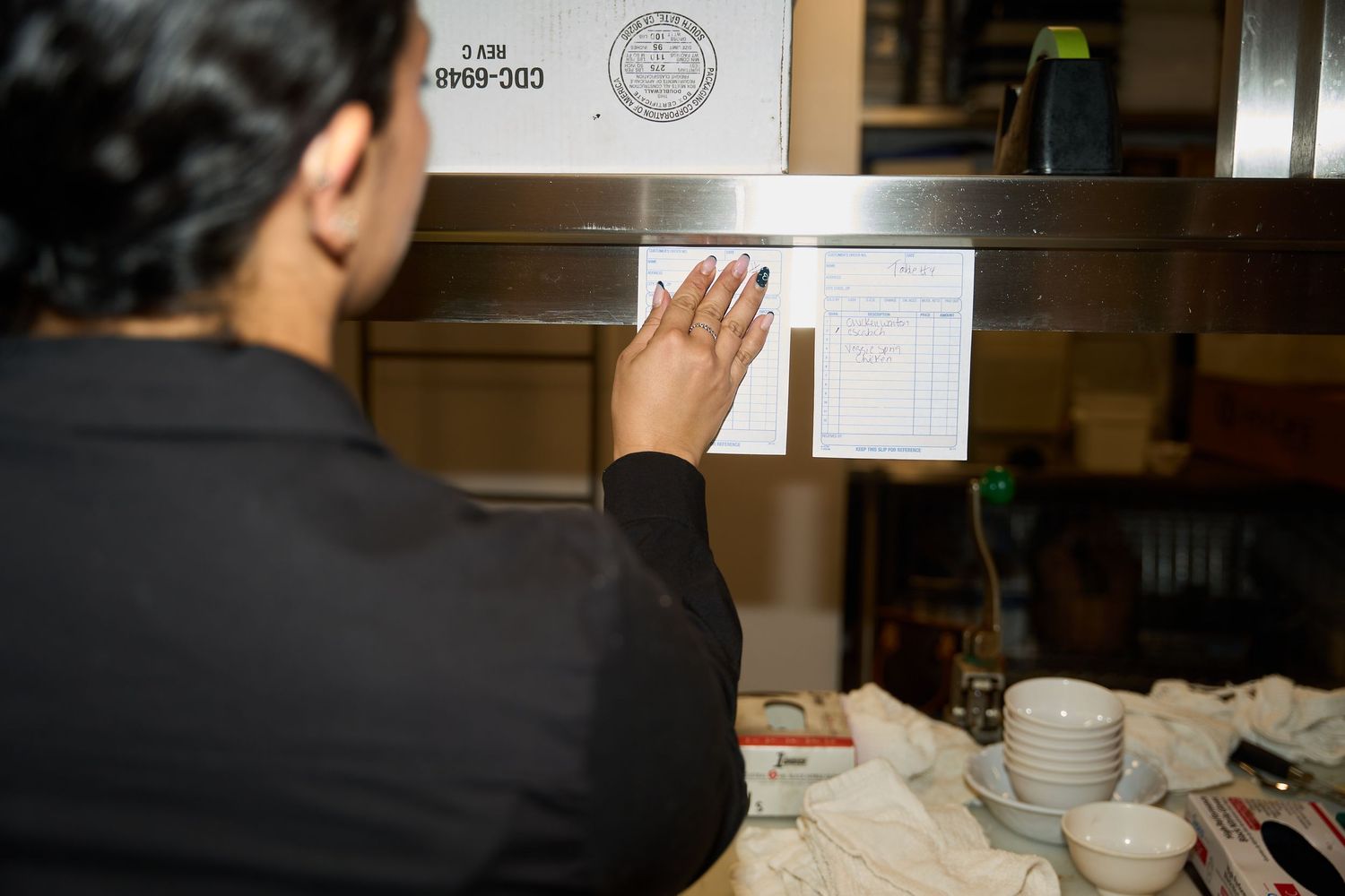 Restaurant worker in black uniform pins an order ticket to a rail in a busy kitchen, with stacked white bowls and plates on the counter.