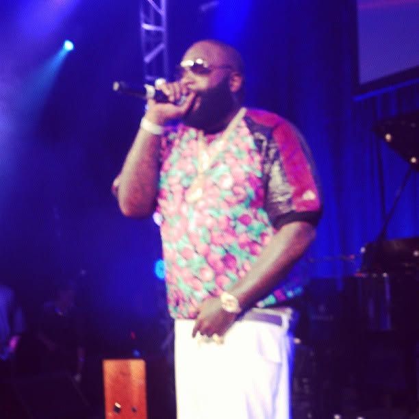 Bearded performer in sunglasses and a colorful floral shirt sings into a microphone on stage under bright blue concert lights.