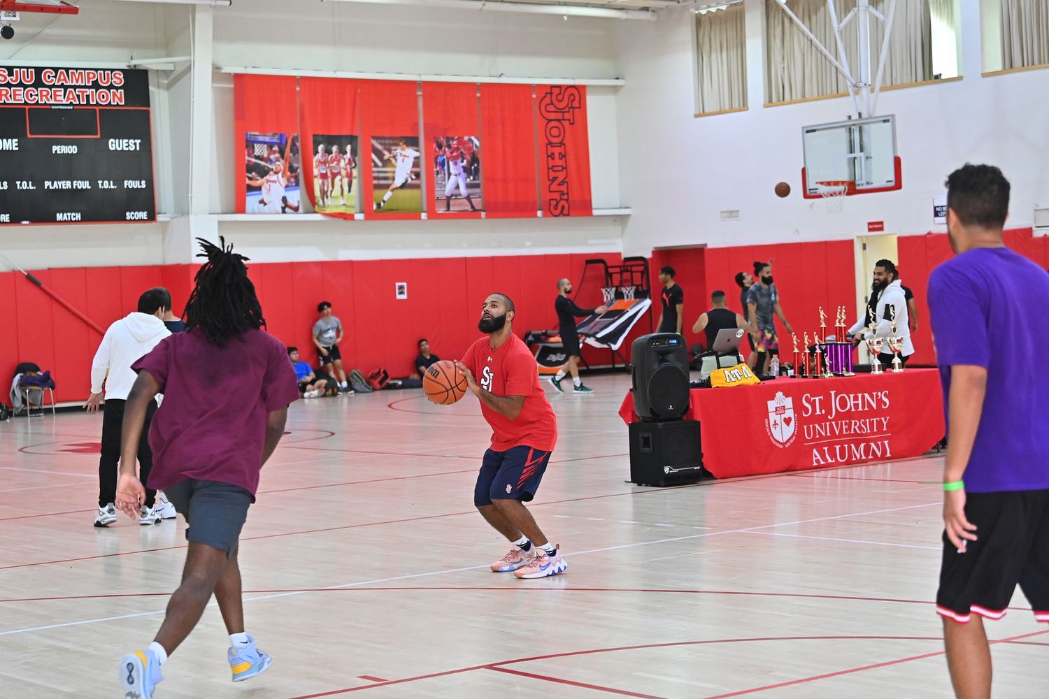 Wide view of a basketball gymnasium with red walls showing multiple players spread across the court during practice.
