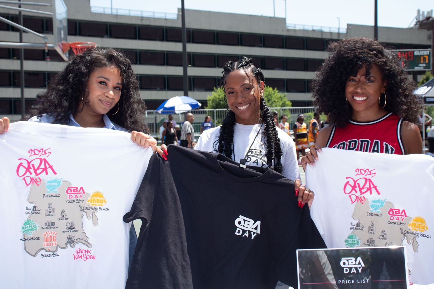 Three people show off t-shirts with colorful artwork designs at an outdoor event.