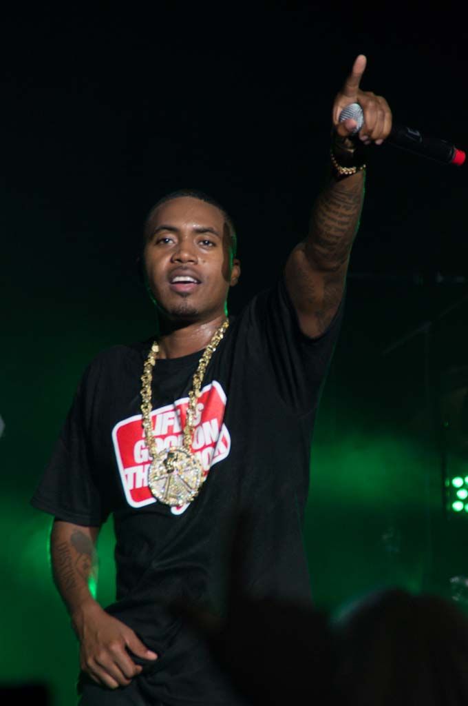 Performer in a black t-shirt and gold chain performing on stage with raised arm against dark background with green lighting.