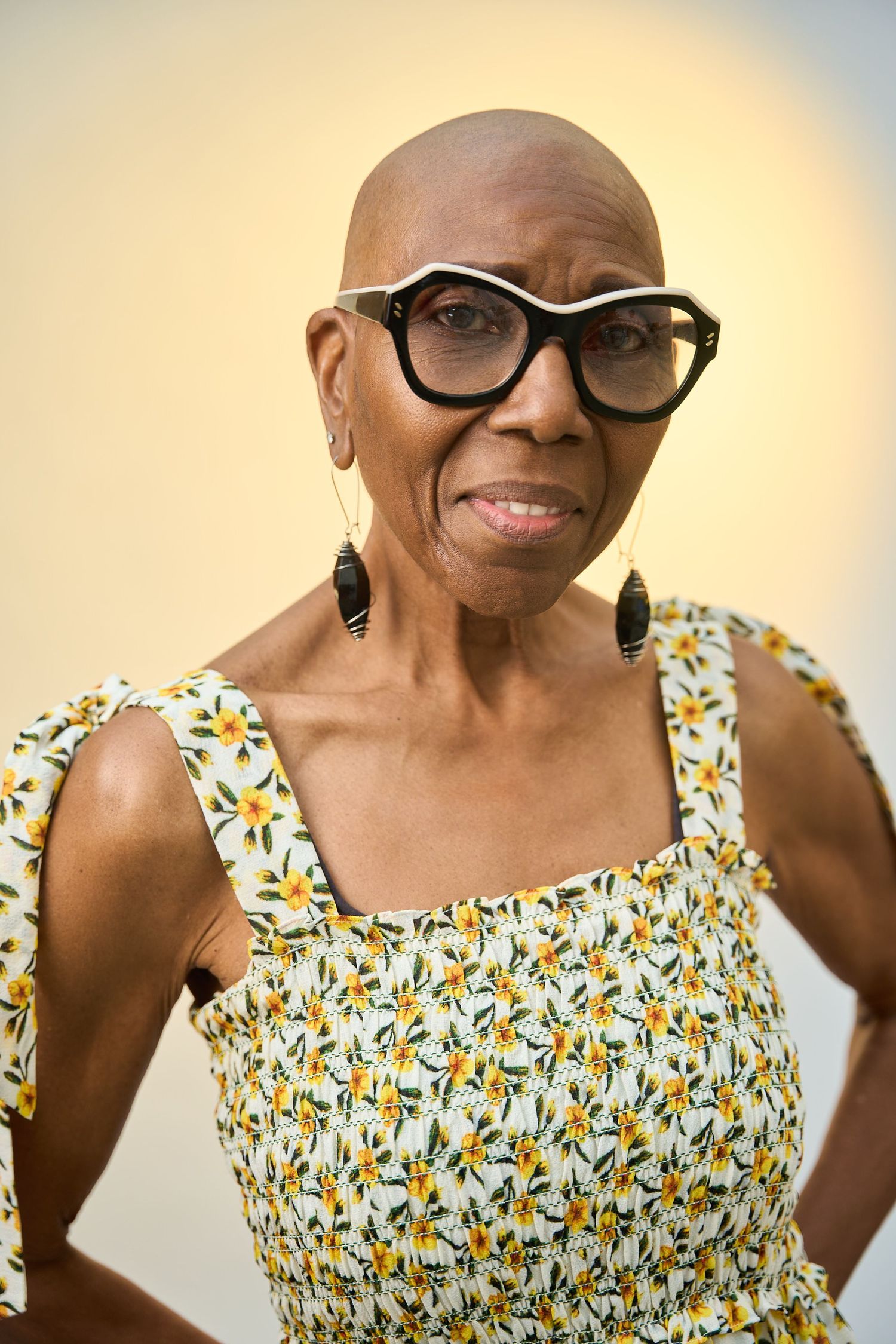 Elderly Black woman with bold glasses and floral sundress poses confidently with hands on hips against neutral background.