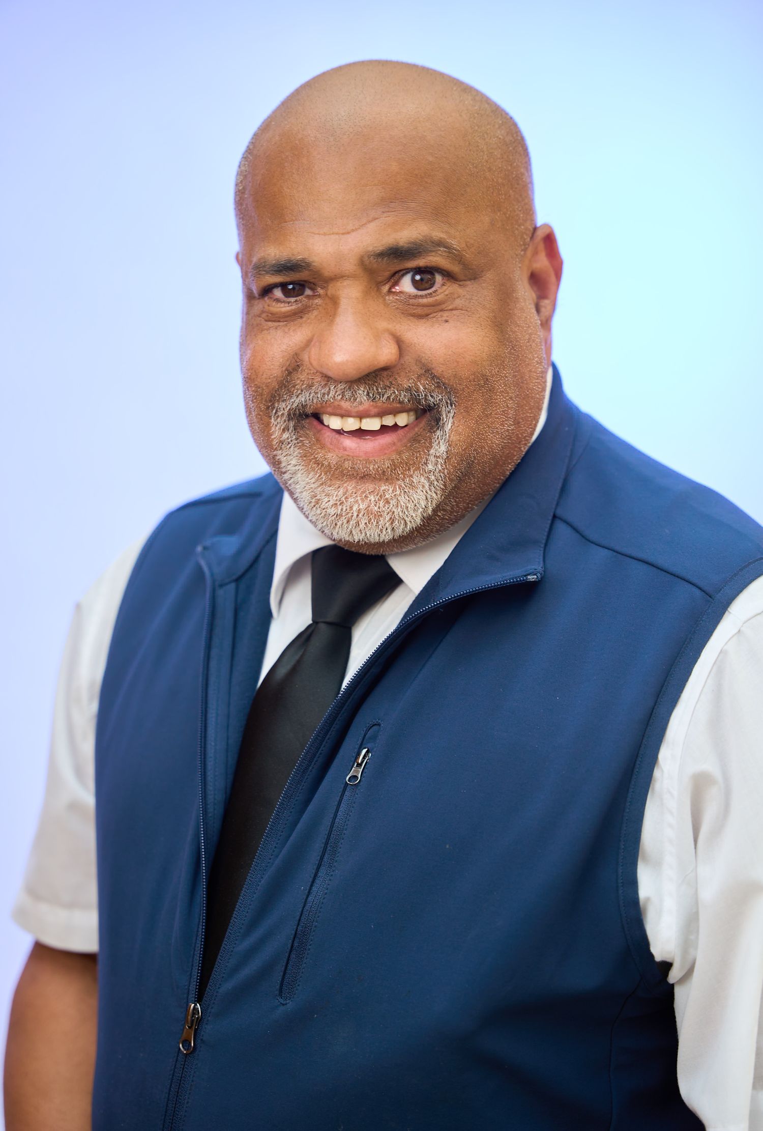 Smiling bald man with gray beard wearing blue vest and black tie against light blue background.