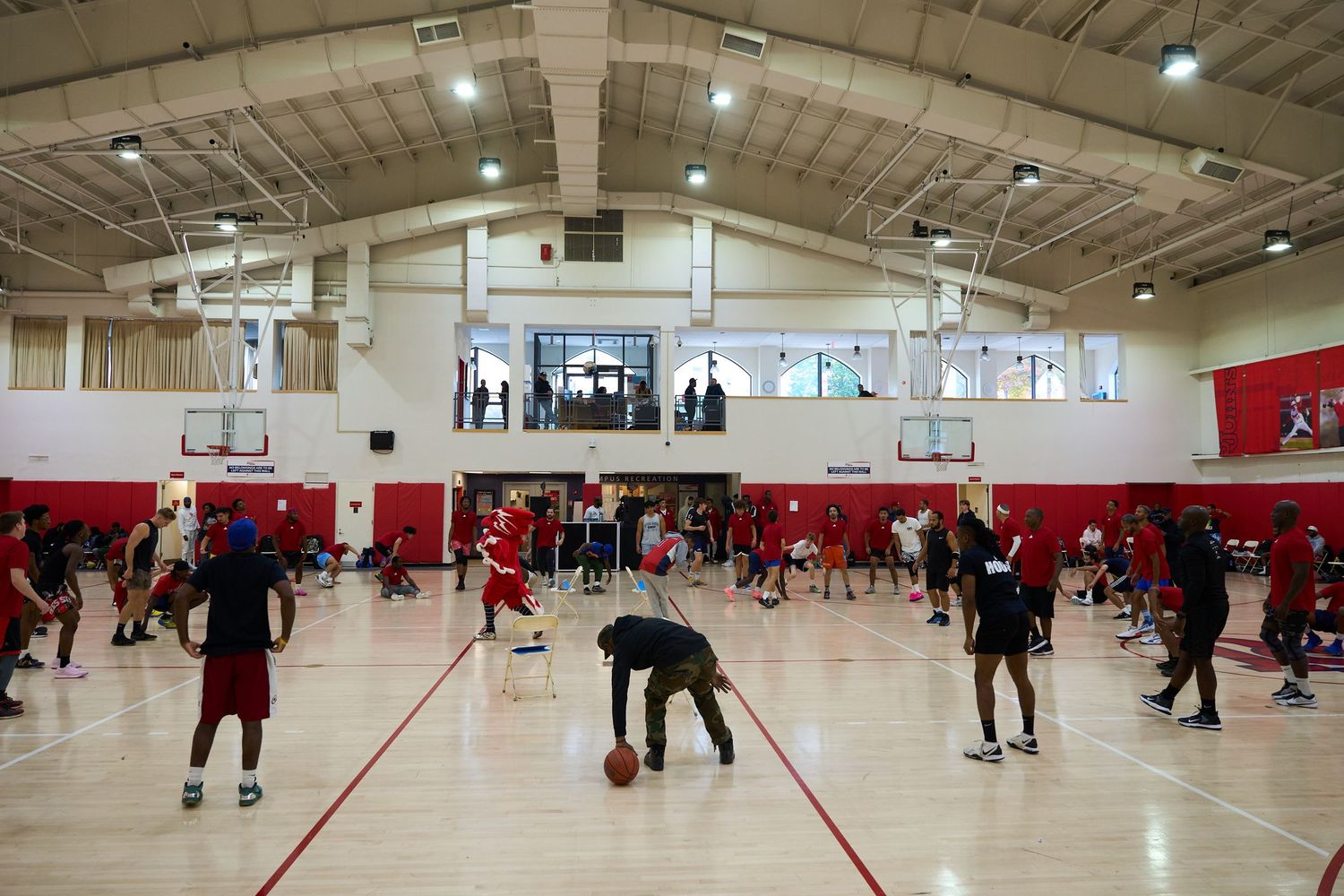 Basketball players practicing drills in a large indoor gymnasium with red walls.