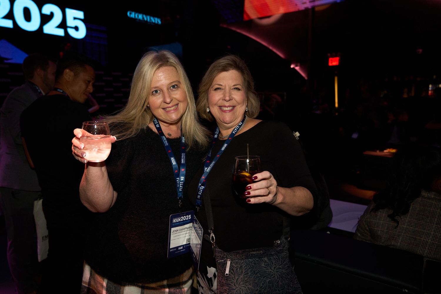 Two attendees enjoying themselves at a nighttime event with lanyards and drinks.