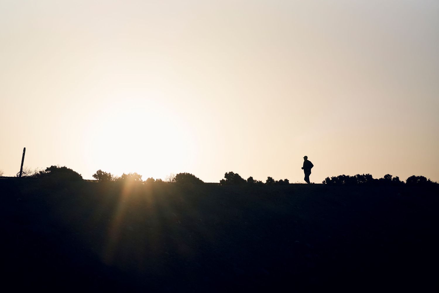 Silhouette of a person walking on a hilltop against a bright sunset sky with rays of light peeking through.