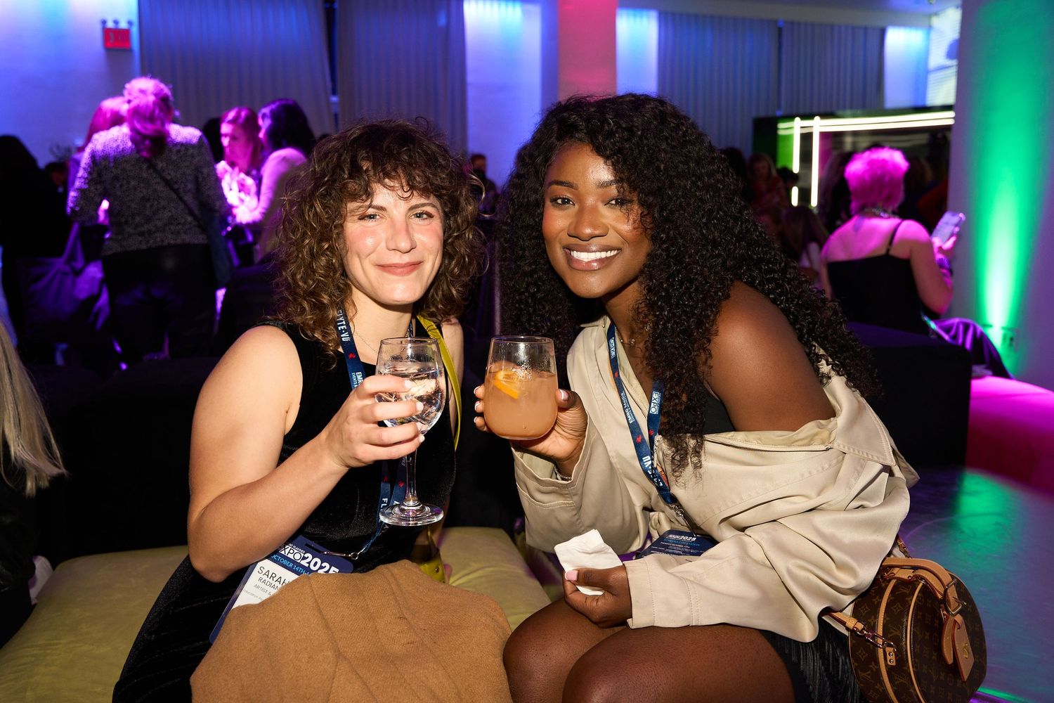 Friends enjoying drinks and socializing at a nightclub with colorful purple and blue lighting effects in the background.