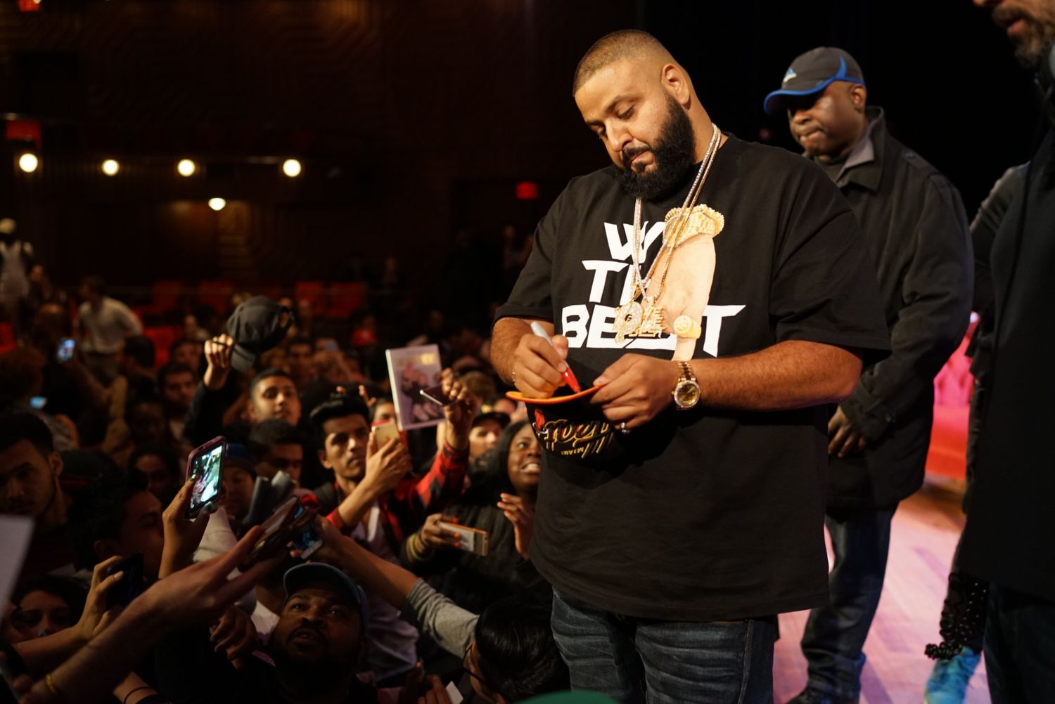 DJ signing a fan's merchandise at a dark nightclub event with crowd in background.