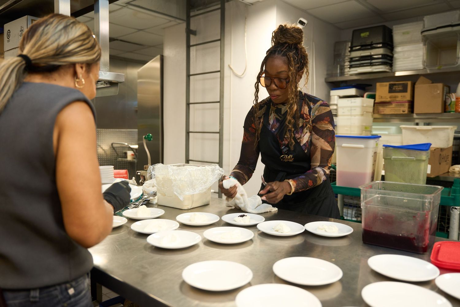 Two chefs prepare and plate food on white dishes lined up along a stainless steel counter in a busy commercial kitchen.
