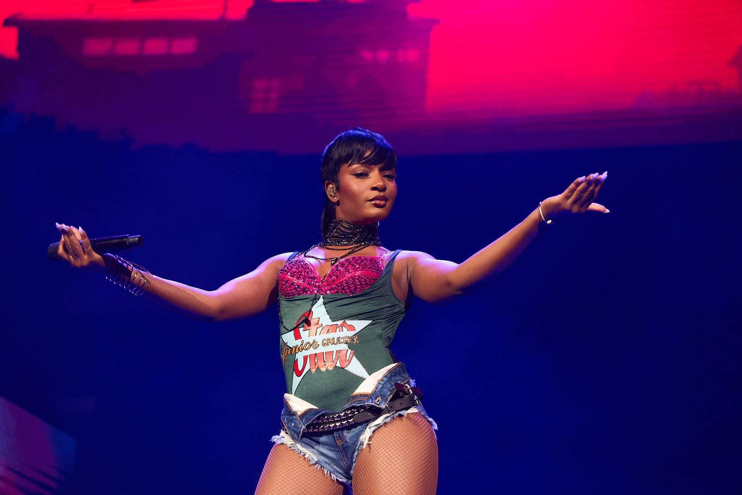 Stage performer in a green and white graphic t-shirt dances energetically under vibrant red and blue stage lighting.