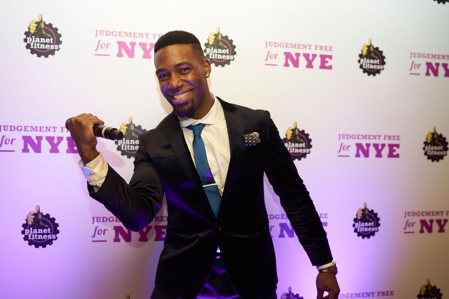 A person in a dark suit and blue tie poses energetically against a backdrop with repeating NYE text and logos.