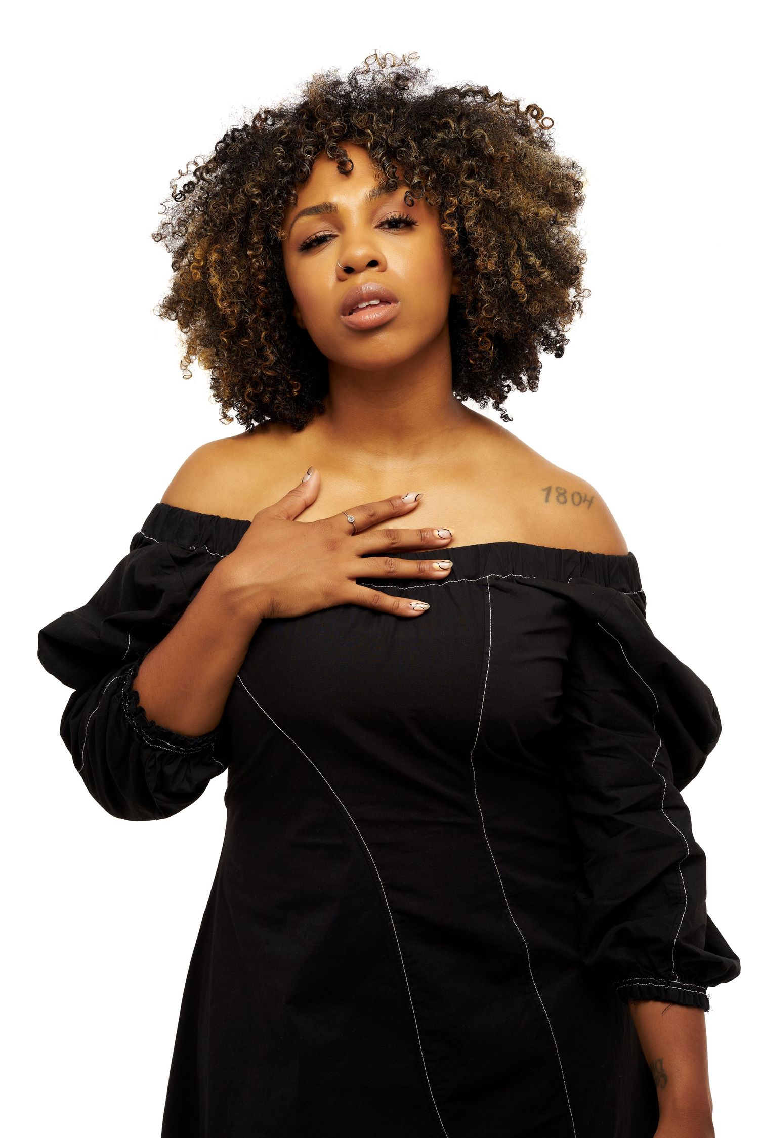 A person with voluminous curly natural hair poses in black off-shoulder dress against white background.