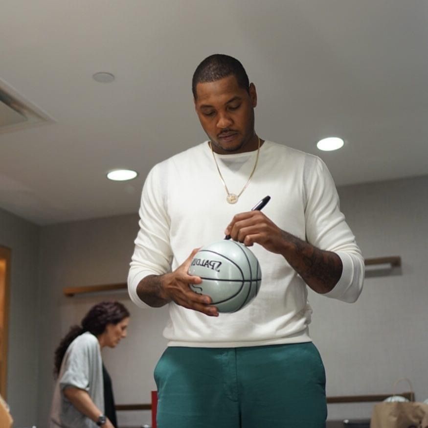 A person in a white sweater autographs a basketball while standing indoors under recessed lighting.