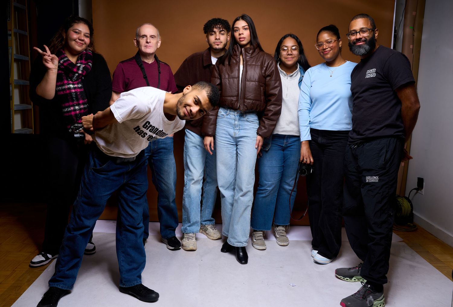 A group of eight people pose together casually in a hallway, with one person playfully leaning forward.
