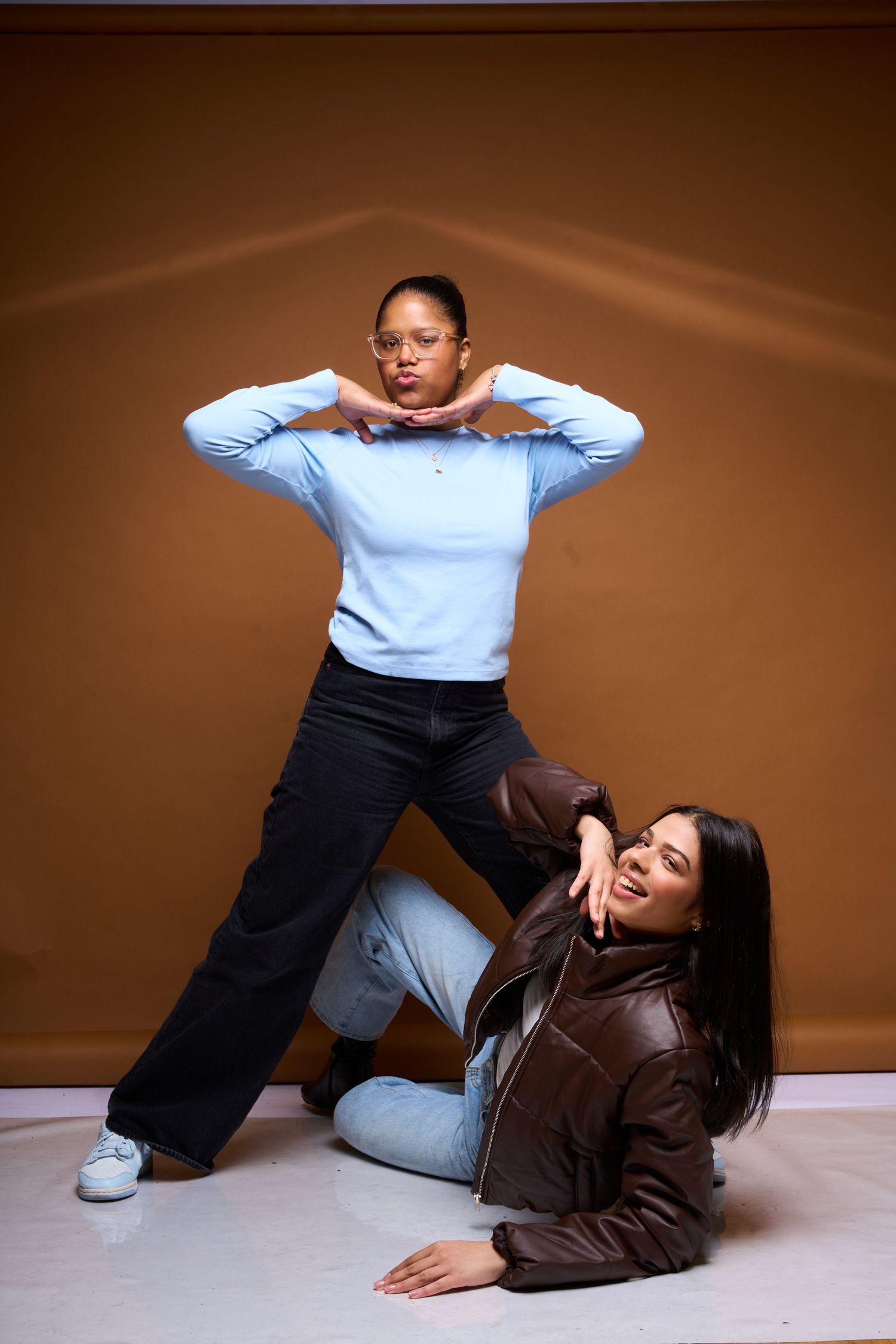 Two people in casual outfits striking dynamic poses against a brown backdrop during a fashion photoshoot.
