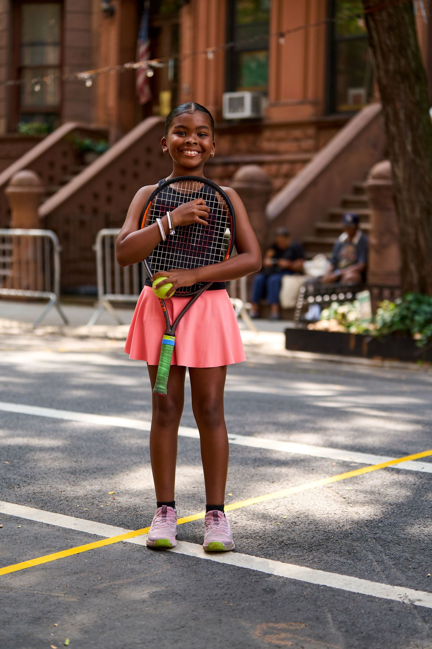 A person wearing a patterned top and pink skirt stands on a city street in front of brownstone buildings.