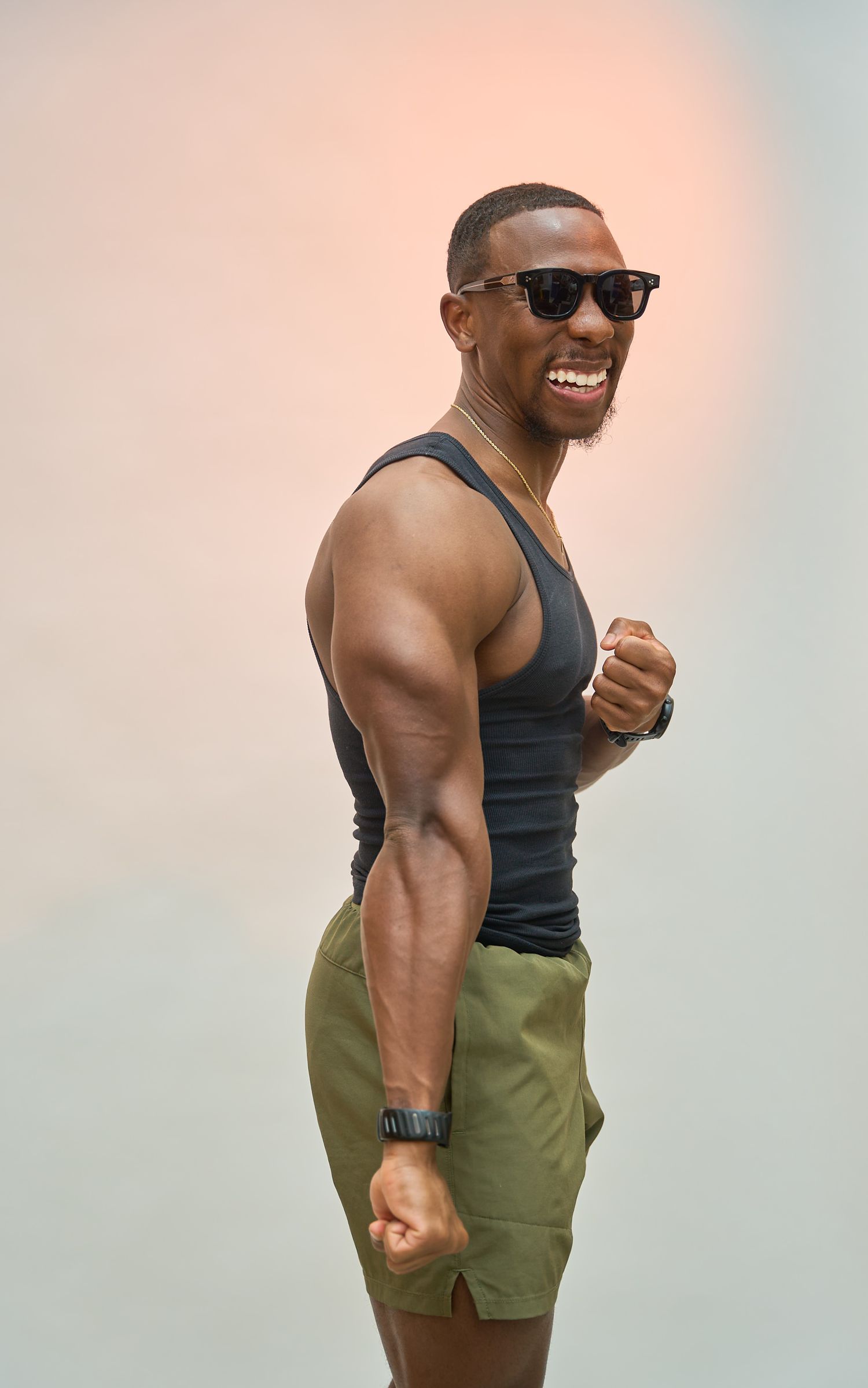 Muscular man in black tank top and green shorts wearing sunglasses, smiling and flexing outdoors.
