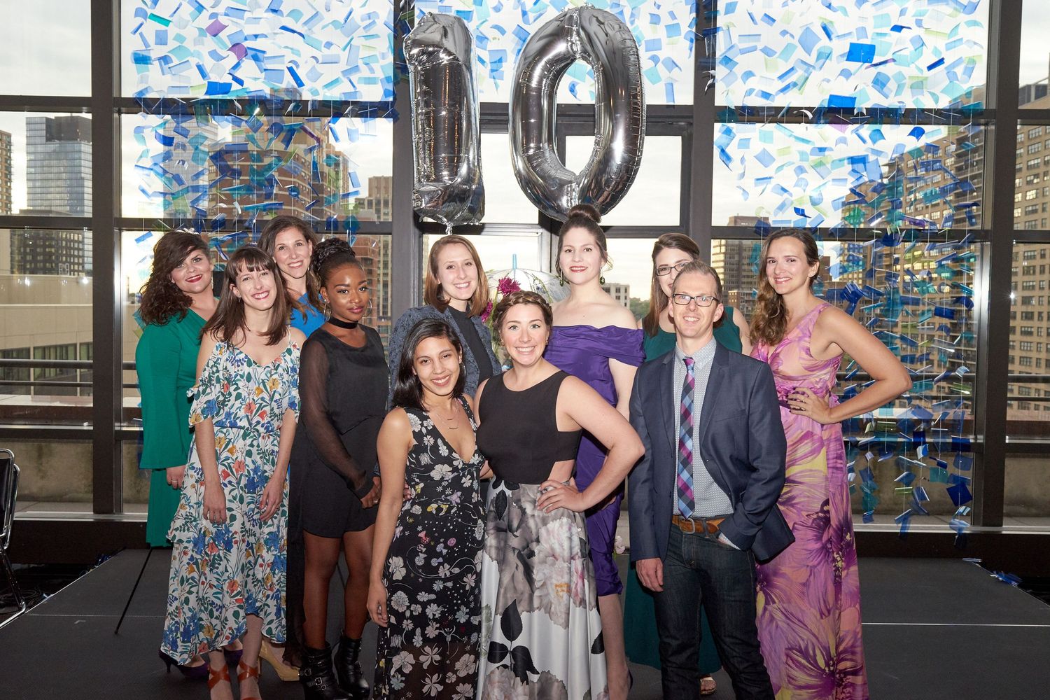 A celebratory group photo with silver number 10 balloons and blue confetti in the background at an event venue.