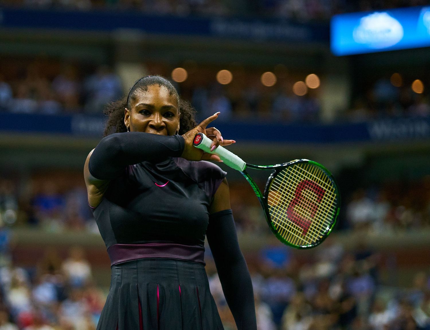 A tennis player serves during match action on a professional court with stadium lights illuminated in background.