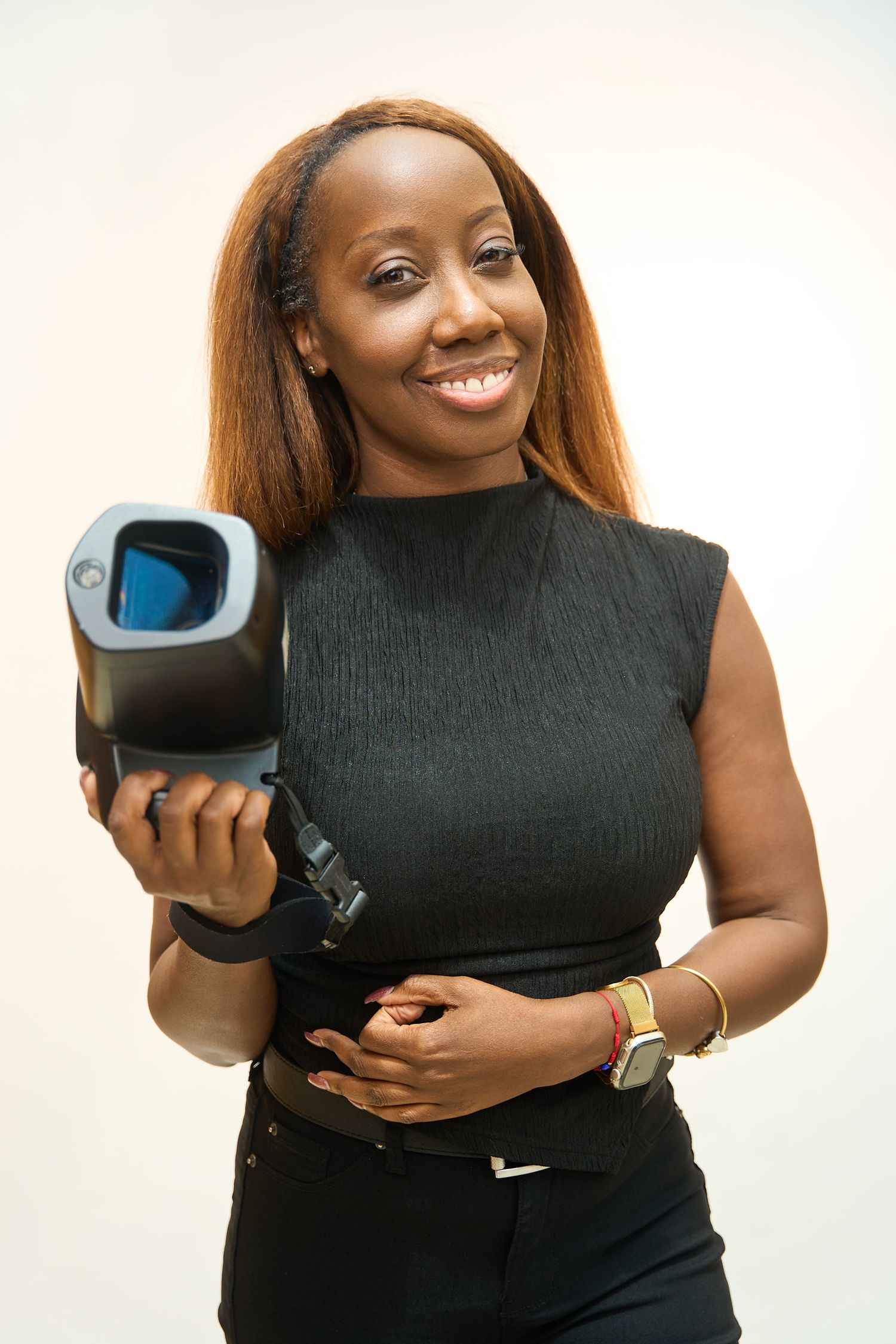 Smiling woman in black sleeveless top holding a video camera, wearing gold jewelry, against a light background.