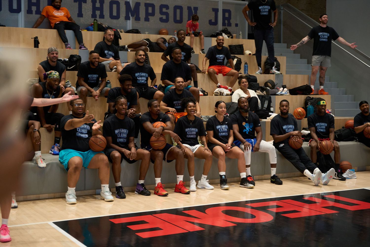 Players sitting on bleachers watching basketball practice in an indoor gym with black flooring.