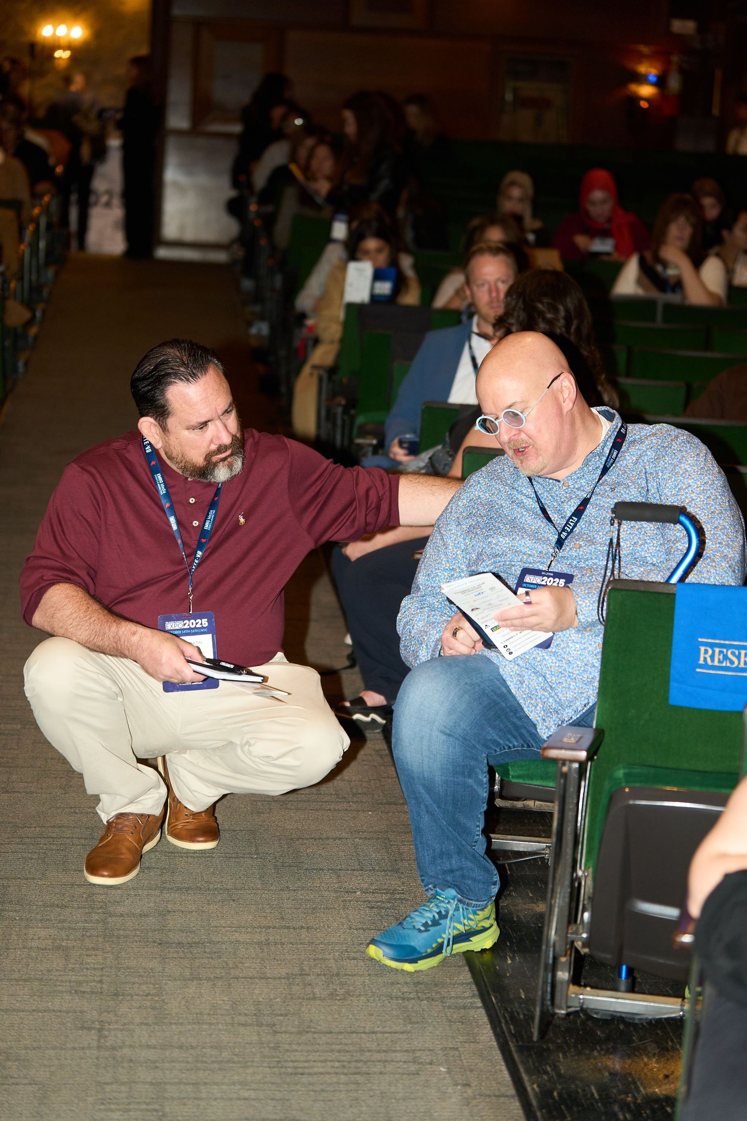 Two people sit on chairs in a dimly lit room while looking down at something in their hands.