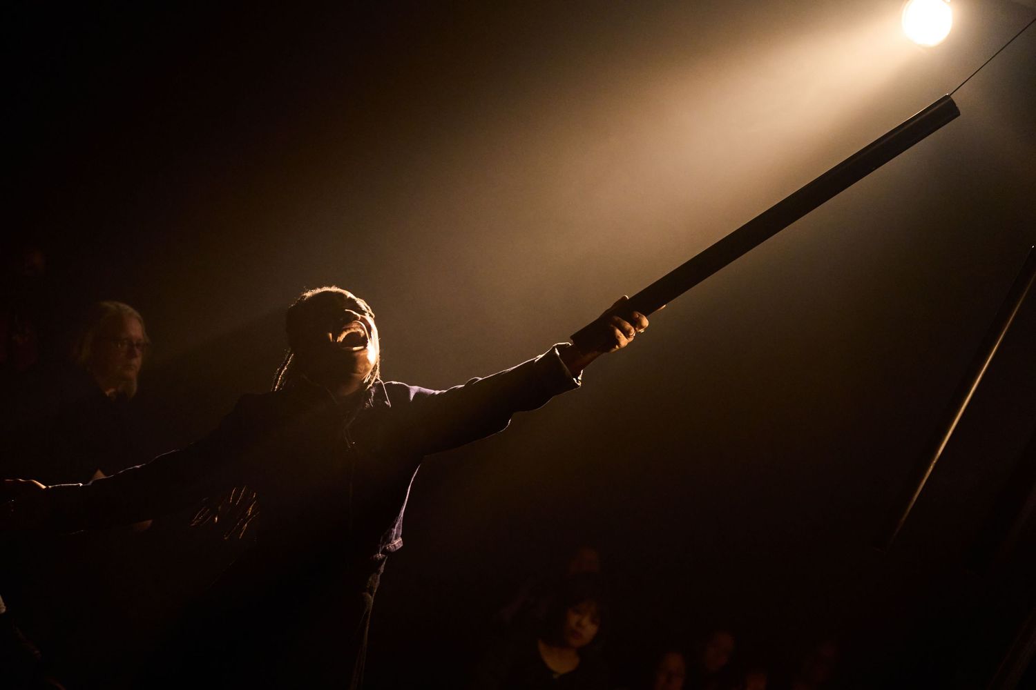 A silhouetted figure holds a long rifle under dramatic stage lighting during a performance.