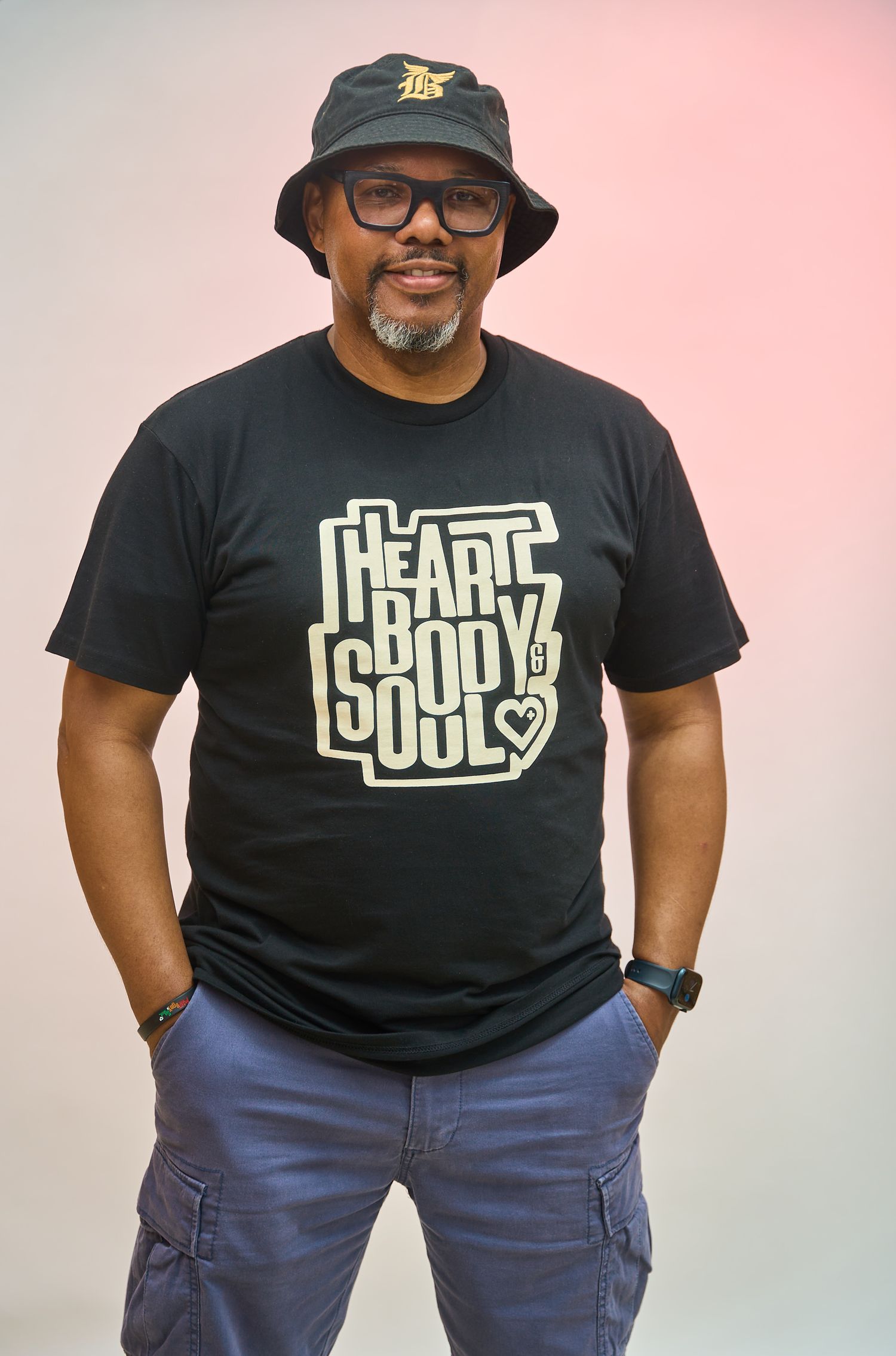 Man smiling in black Heart Body Soul t-shirt and bucket hat, posing confidently with hands in pockets.