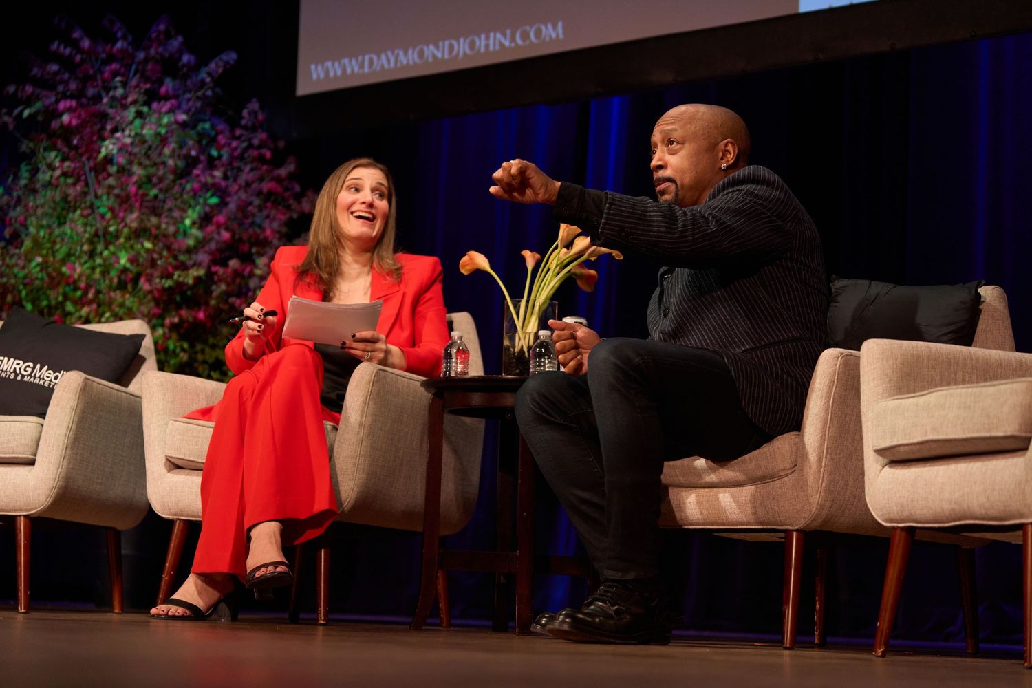 Two people engaged in an animated discussion while seated on stage at a professional event.
