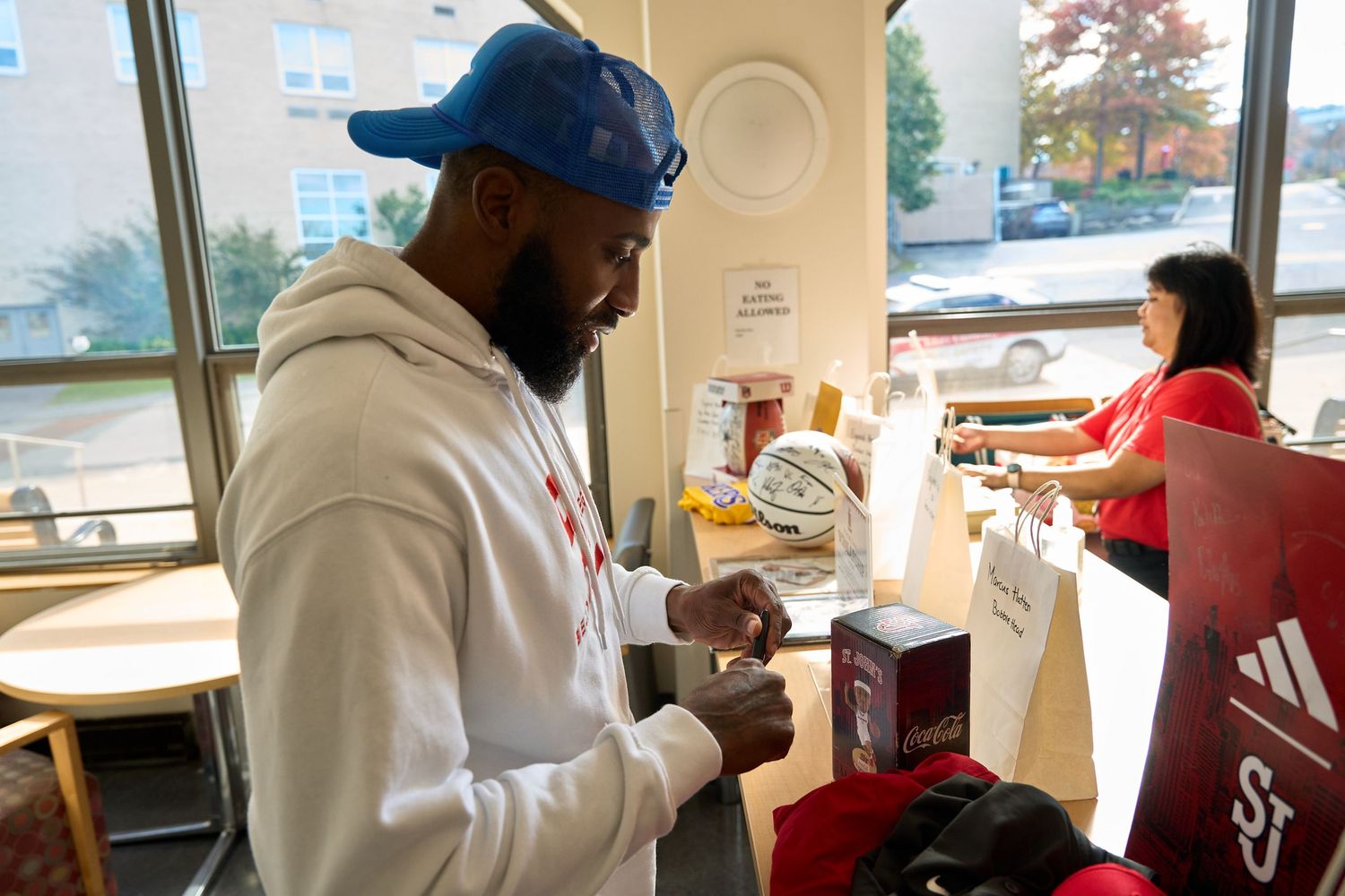 A person in a blue cap and white hoodie examines items at a table in a bright indoor space.