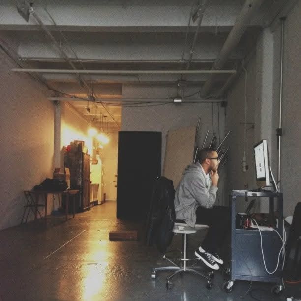 Man in a gray hoodie and glasses sits on a stool, hand on chin, studying a desktop computer in a dim industrial loft studio.