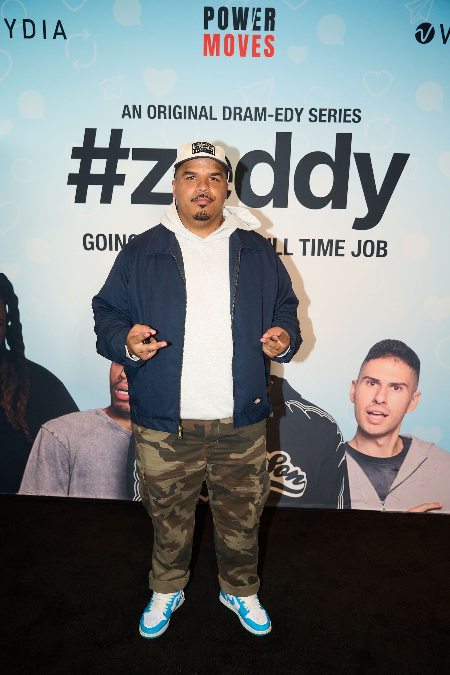 Man in camo pants and blue sneakers poses at #Zeddy dram-edy series premiere event backdrop.