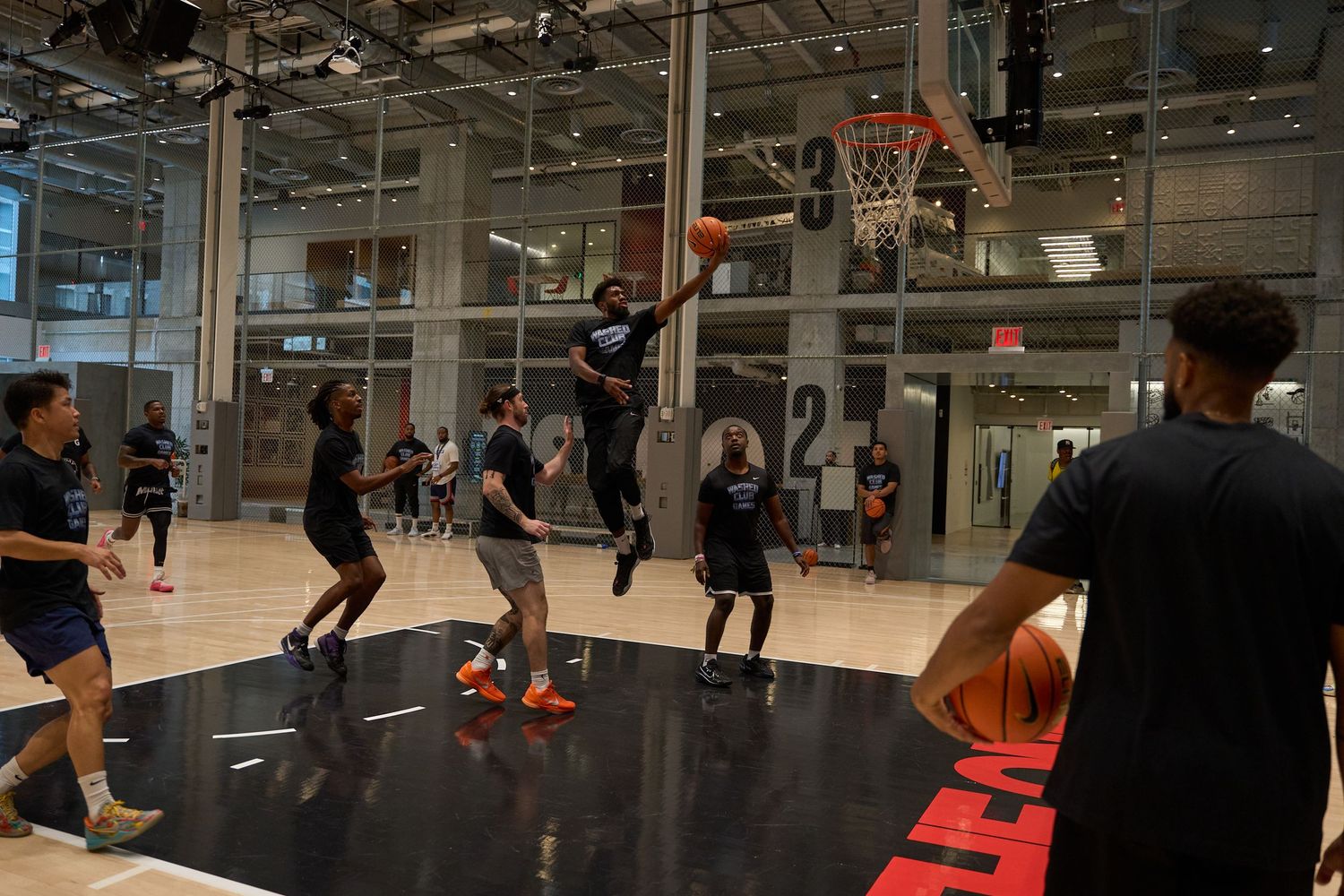 Athletes play basketball on an indoor court with glass walls and modern lighting fixtures overhead.