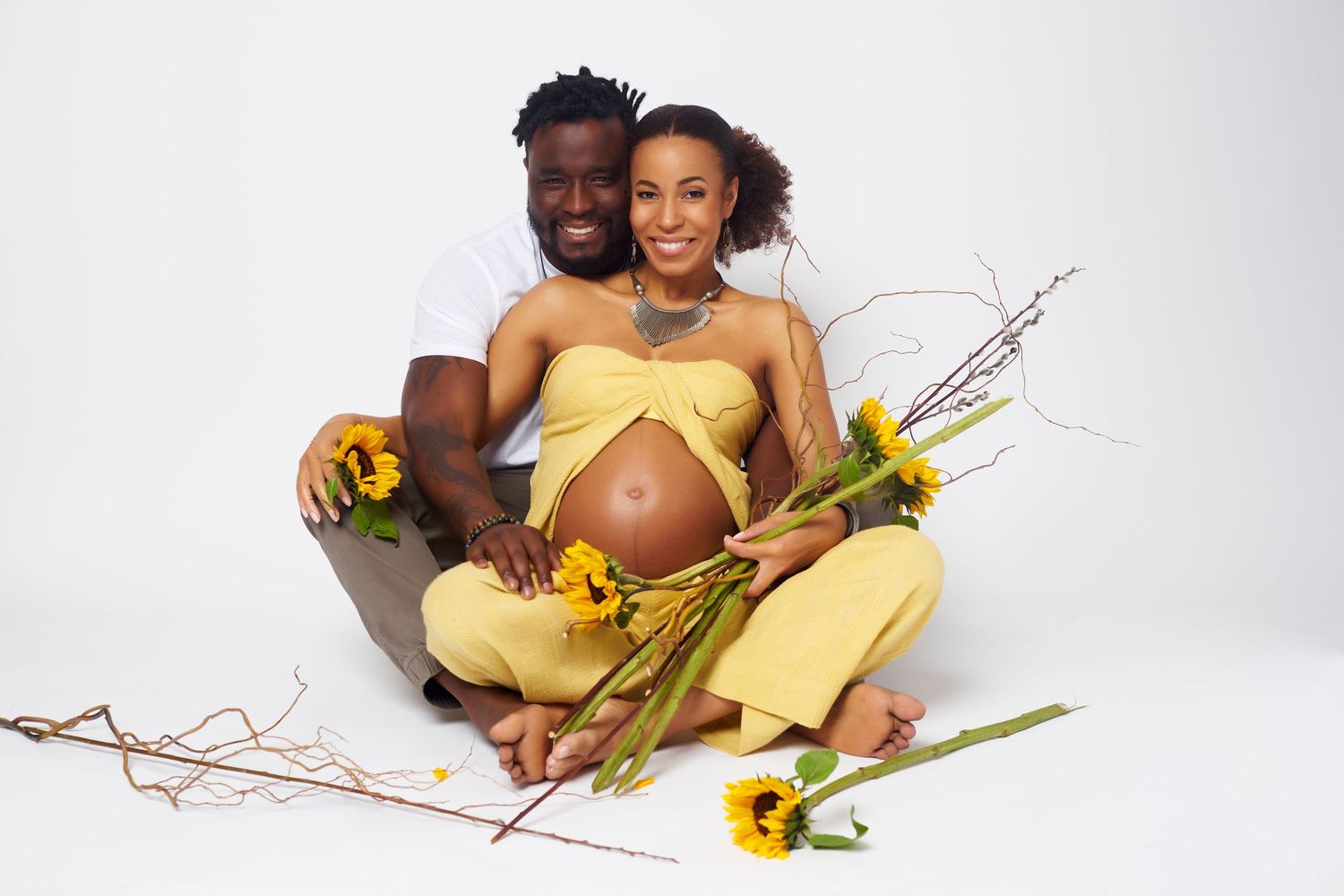 A couple in yellow outfits sits together with sunflowers and branches in a bright studio setting.