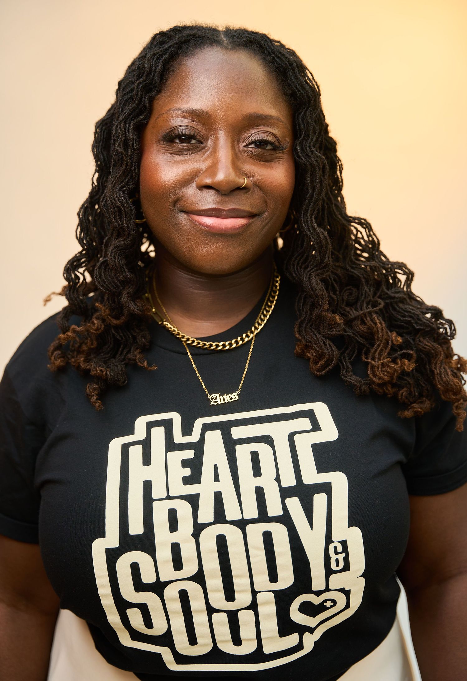 Woman smiling in black Heart Body Soul t-shirt with gold necklace and earrings, wearing locs hairstyle.