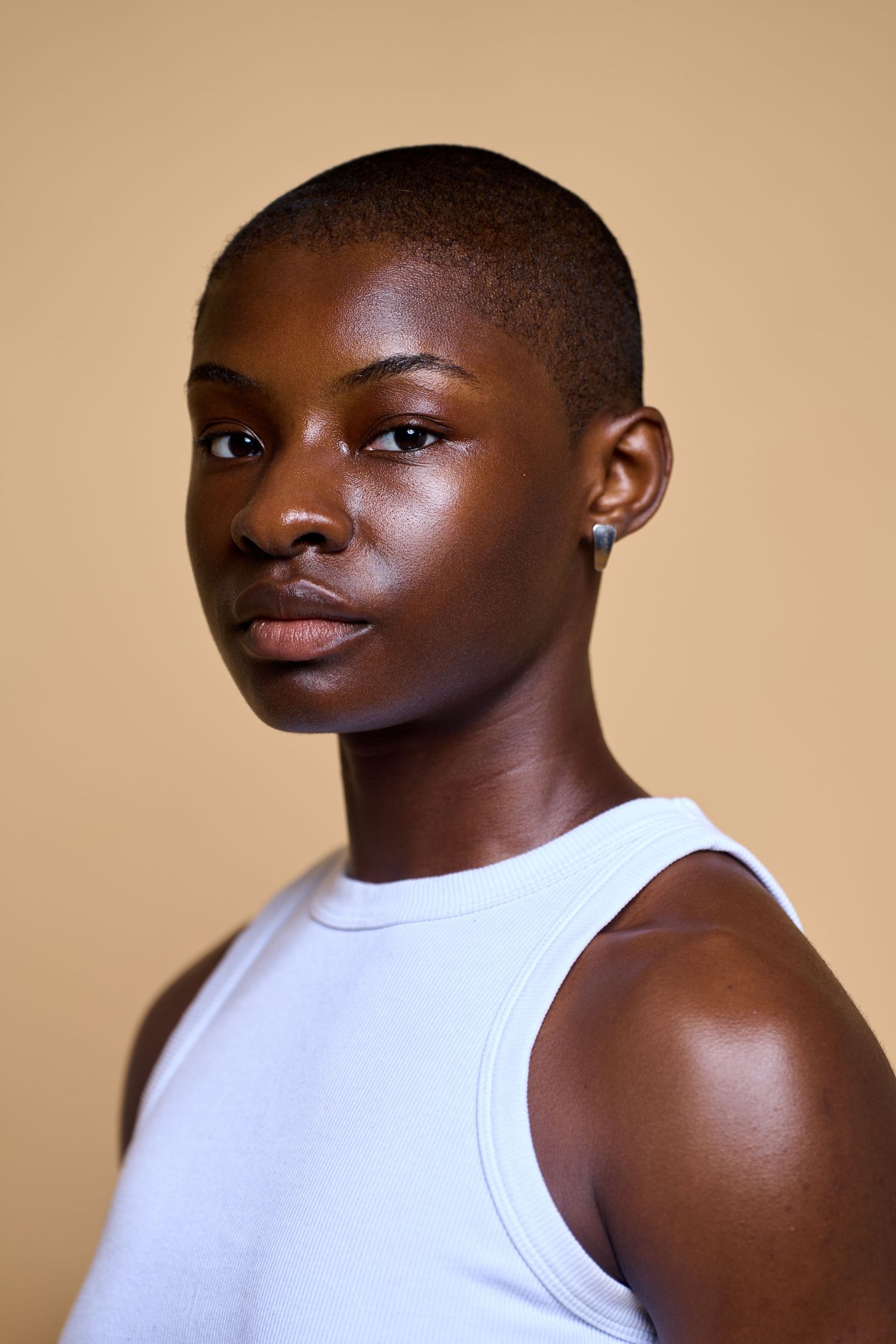 Portrait against beige background showing close up of person in white sleeveless top with short hair and natural glowing skin.