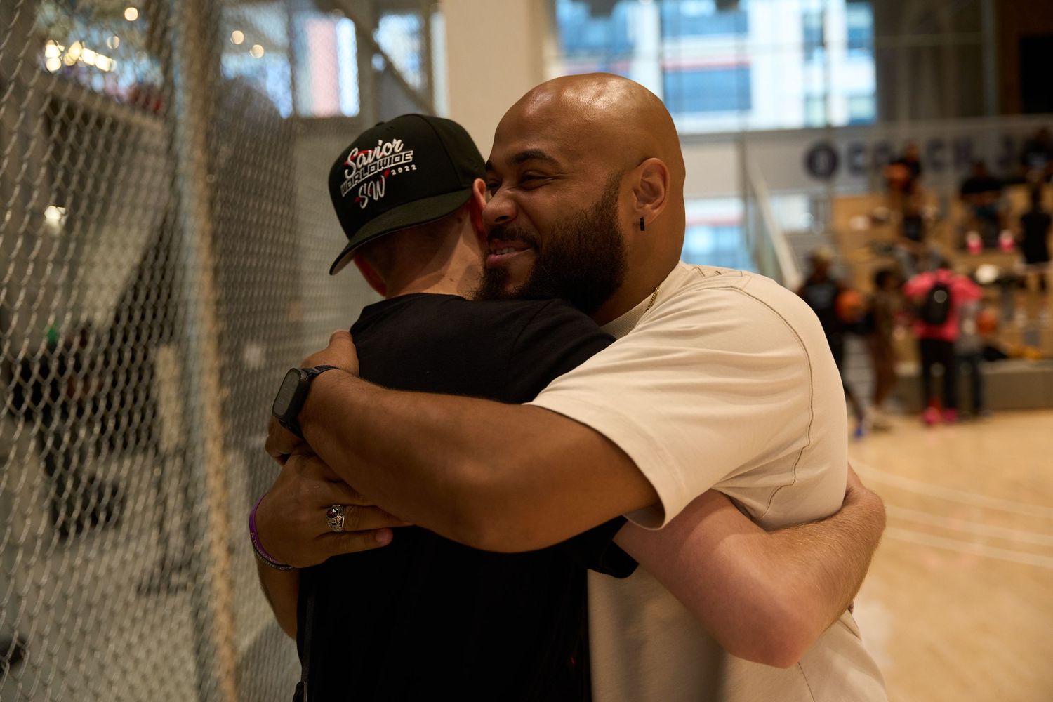Two people share a warm embrace in a gym or indoor sports facility with a chain link fence visible in the background.