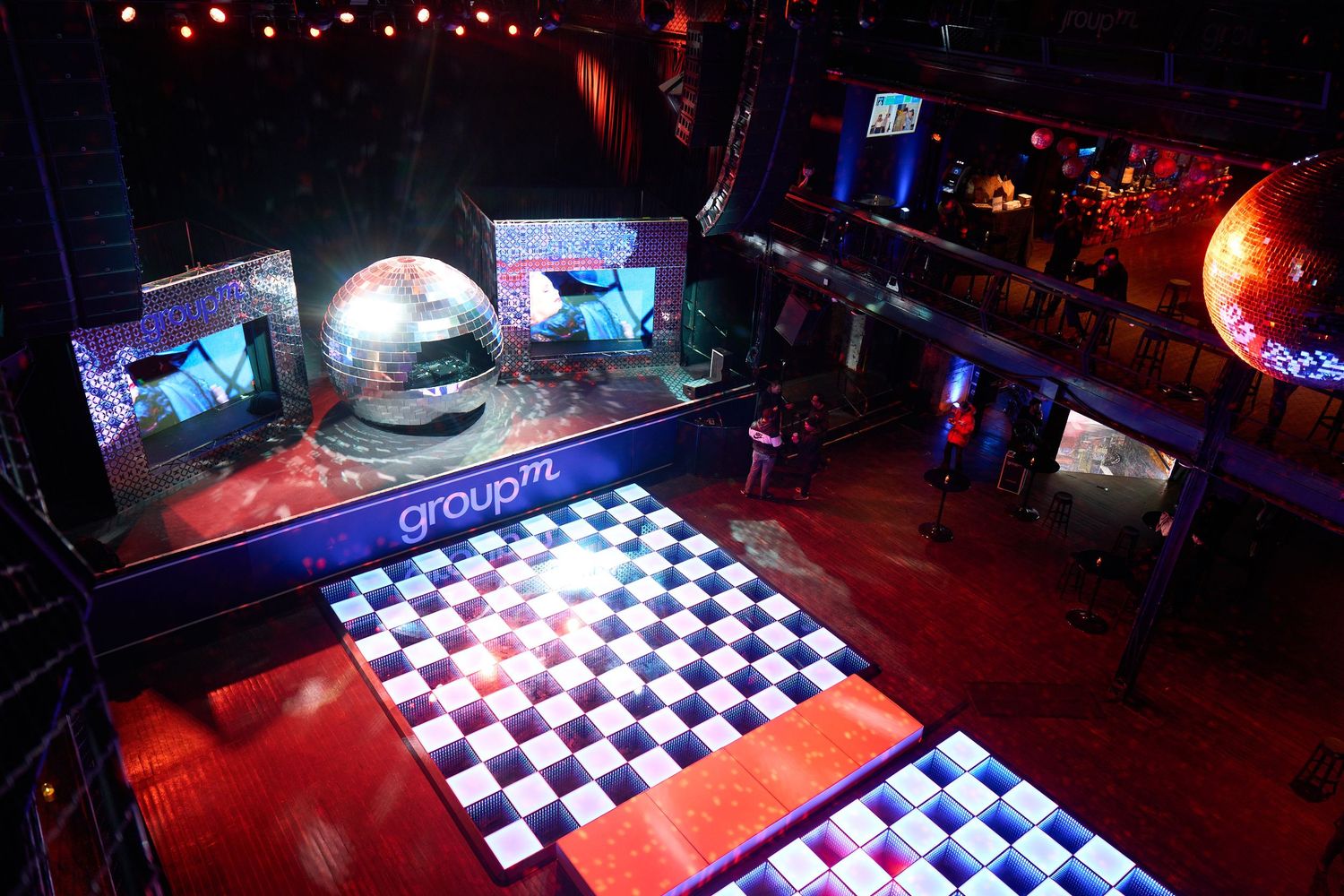 Nightclub interior with illuminated checkered dance floor, disco ball, and multiple screens displaying colorful visuals.