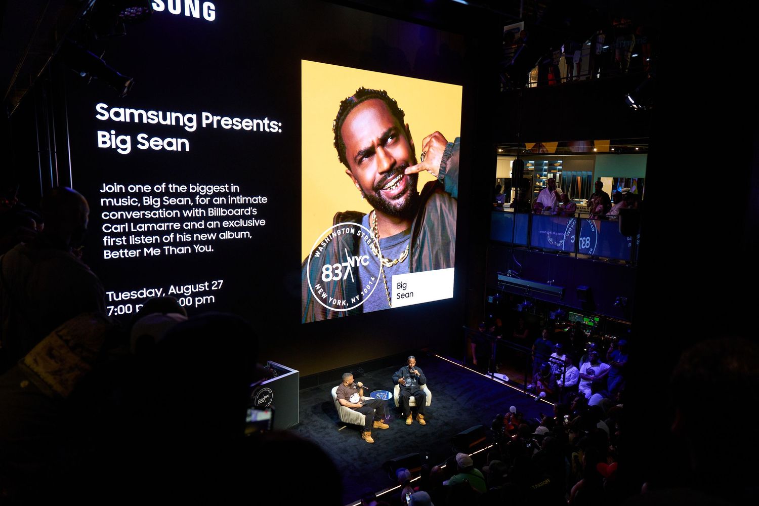 Samsung presentation event at a large indoor venue with digital displays and stage lighting illuminating a dark auditorium.