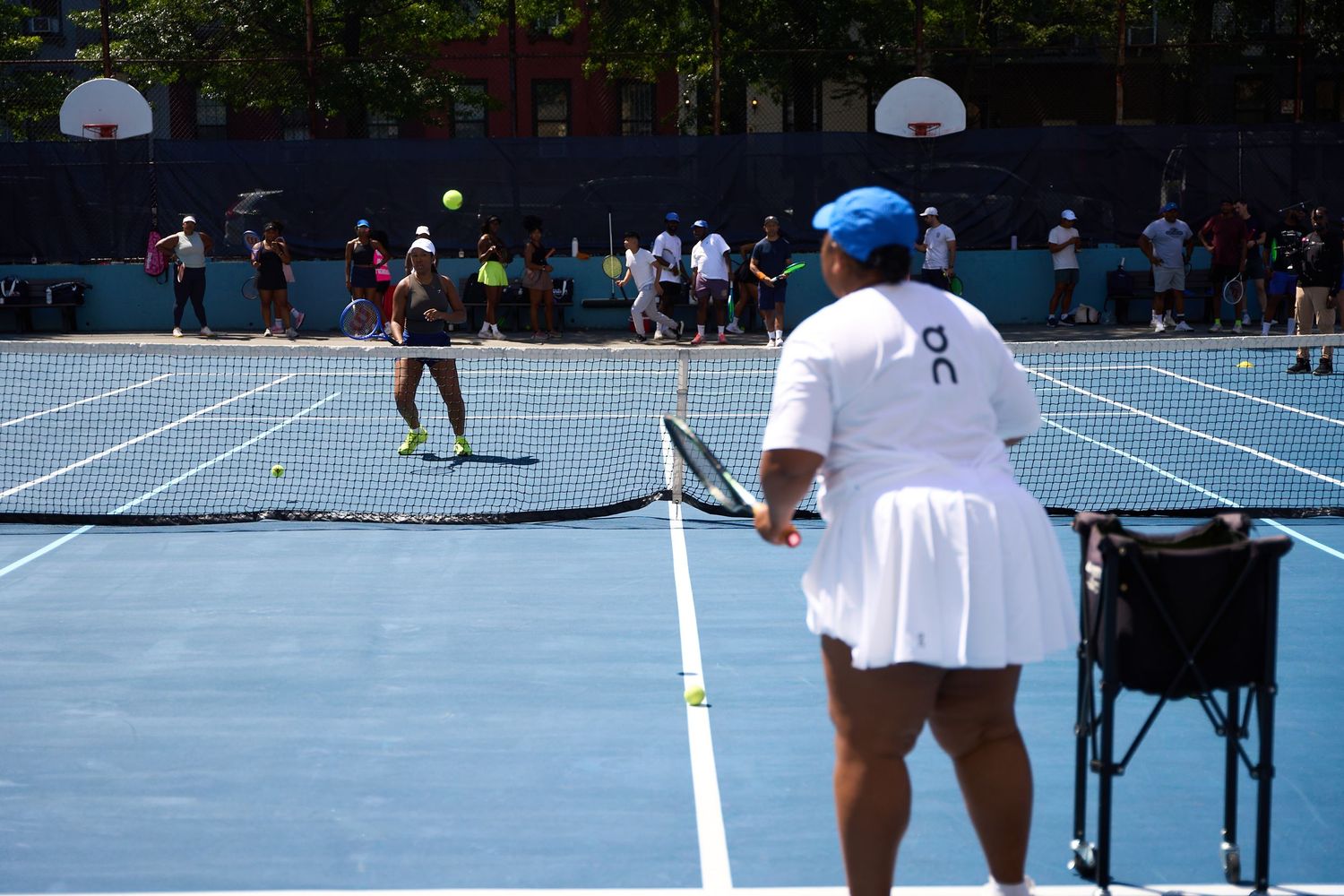 Tennis players and spectators gather on a blue outdoor court during a sunny day.