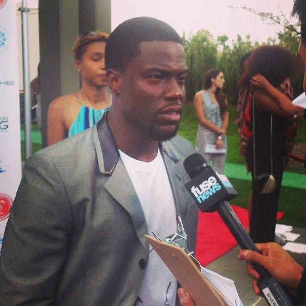 Man in a gray blazer and white t-shirt speaks into a fuse news microphone at an outdoor red carpet event with guests in the background.