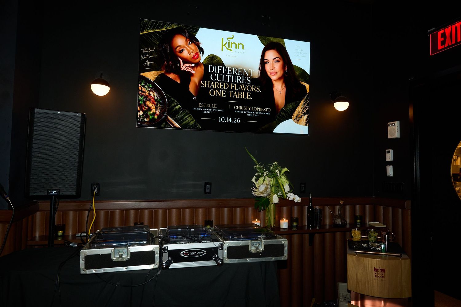 Dimly lit event space with a wall-mounted screen showing a Kinn promotional ad, DJ equipment cases below, and fresh flowers on a table.