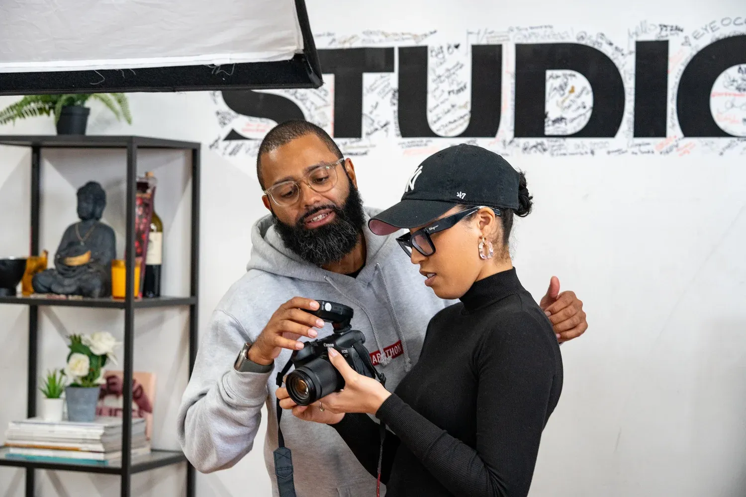 Two photographers review images on a camera display in a studio setting with the word STUDIO visible on the wall.