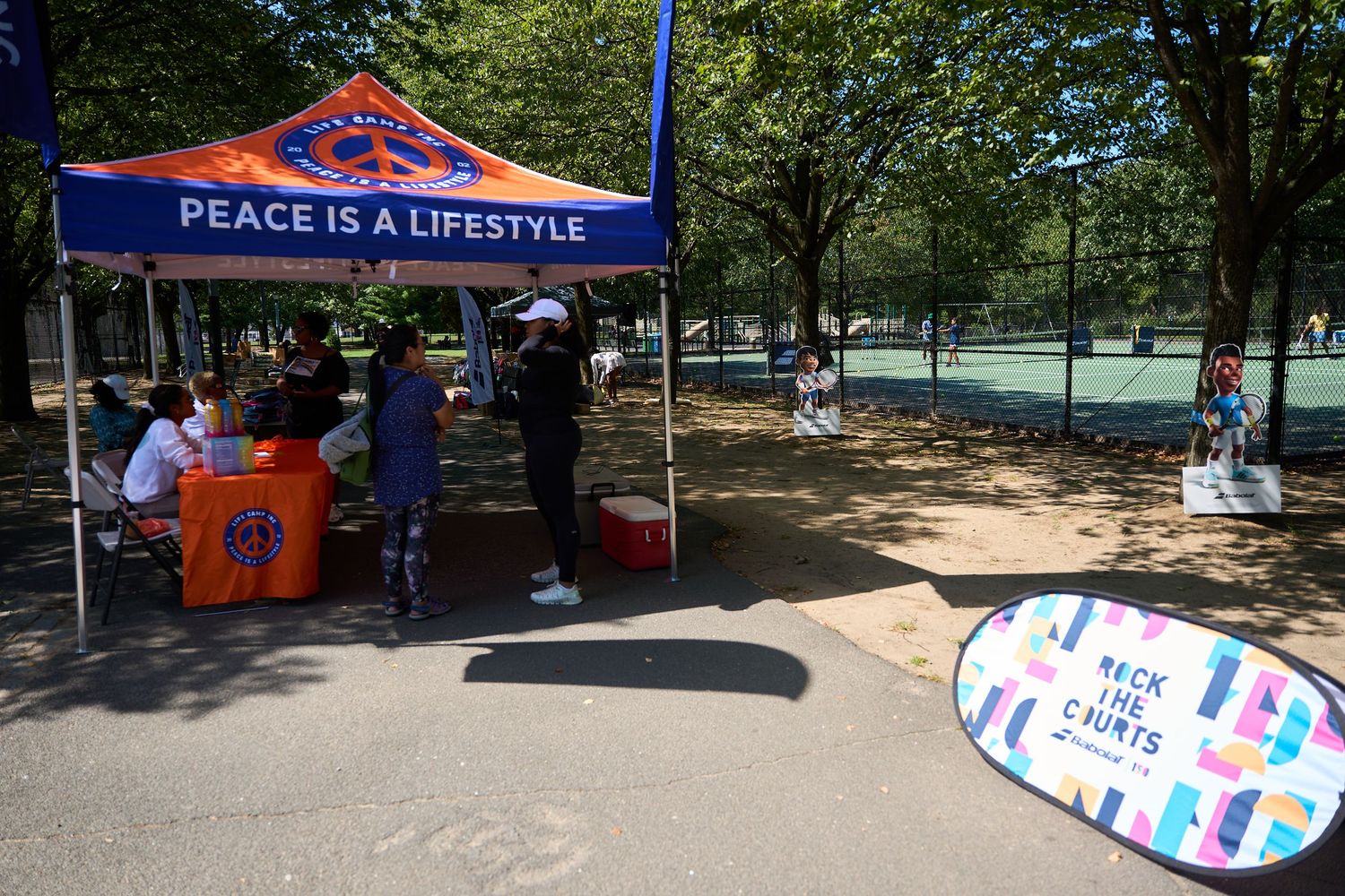 A tent with 'Peace is a Lifestyle' banner at an outdoor event with tennis courts visible in the background.