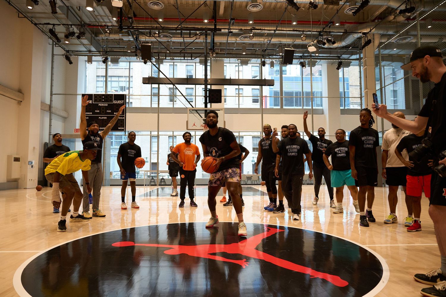 Players gather around a bright red circle on a shiny gymnasium floor during an indoor basketball practice session.