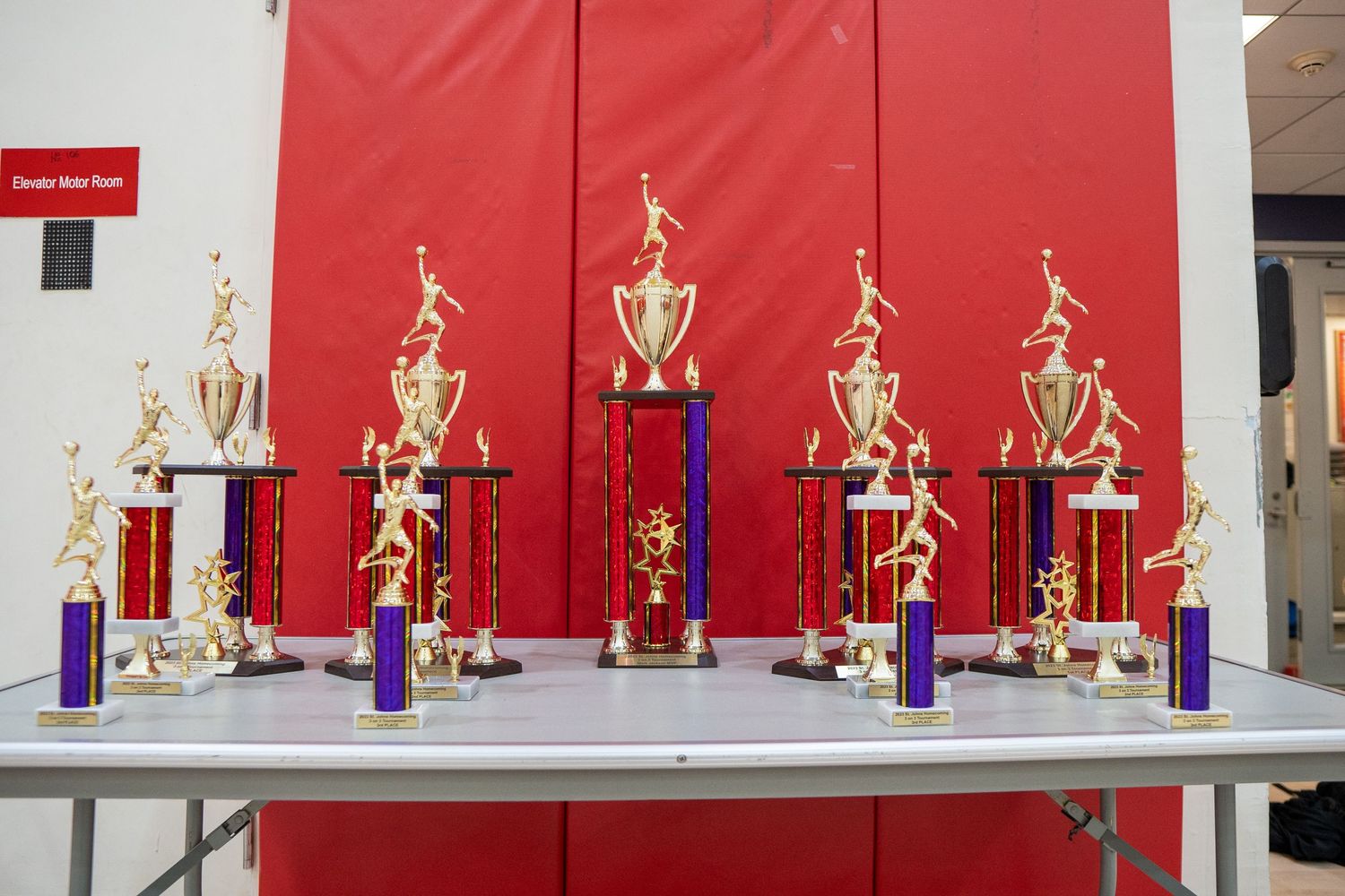 Five tall gold basketball trophies with purple columns stand in a row on a gray table against a red background.