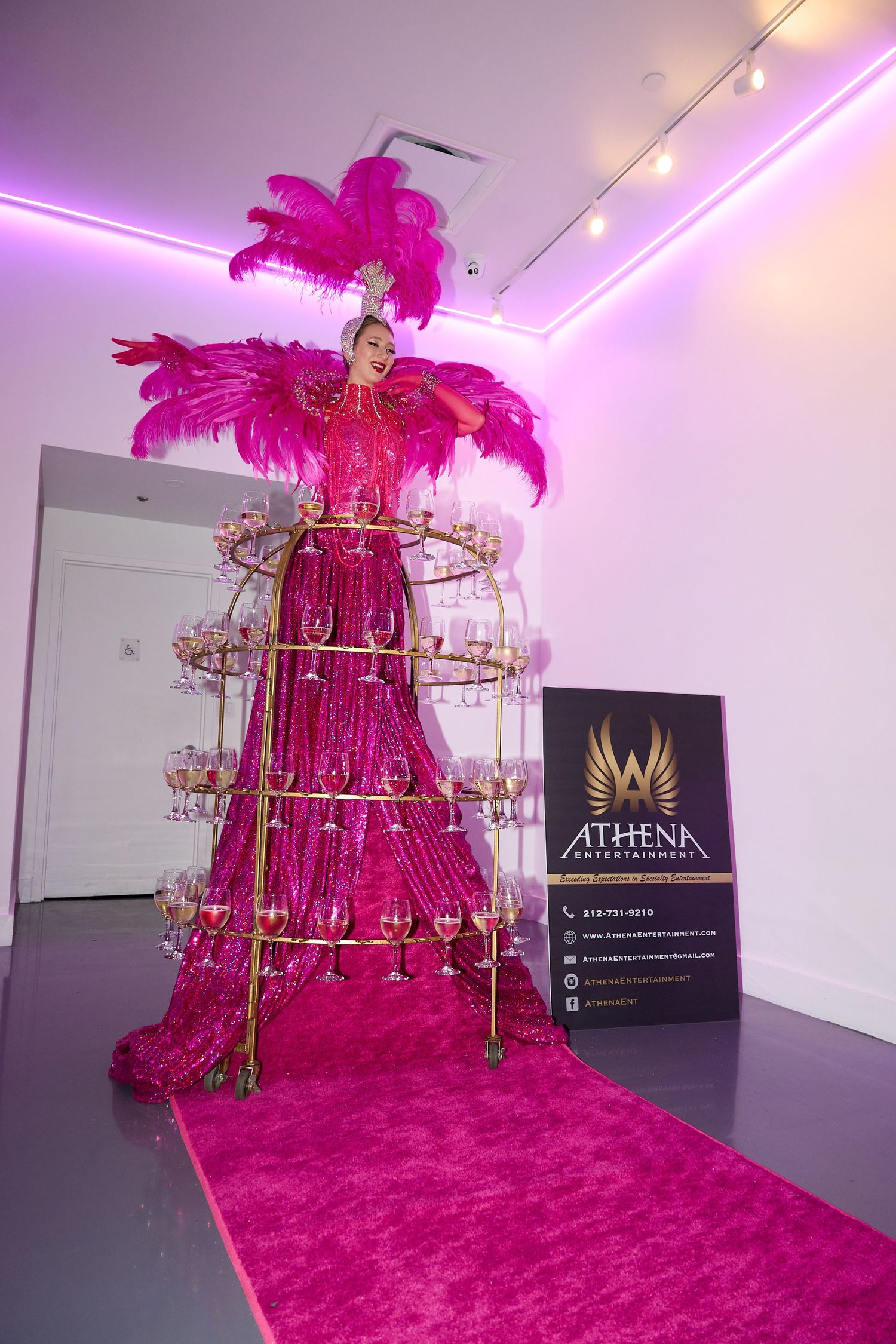 A dramatic pink feathered display tower with champagne stands on a pink carpet under purple lighting at an event.