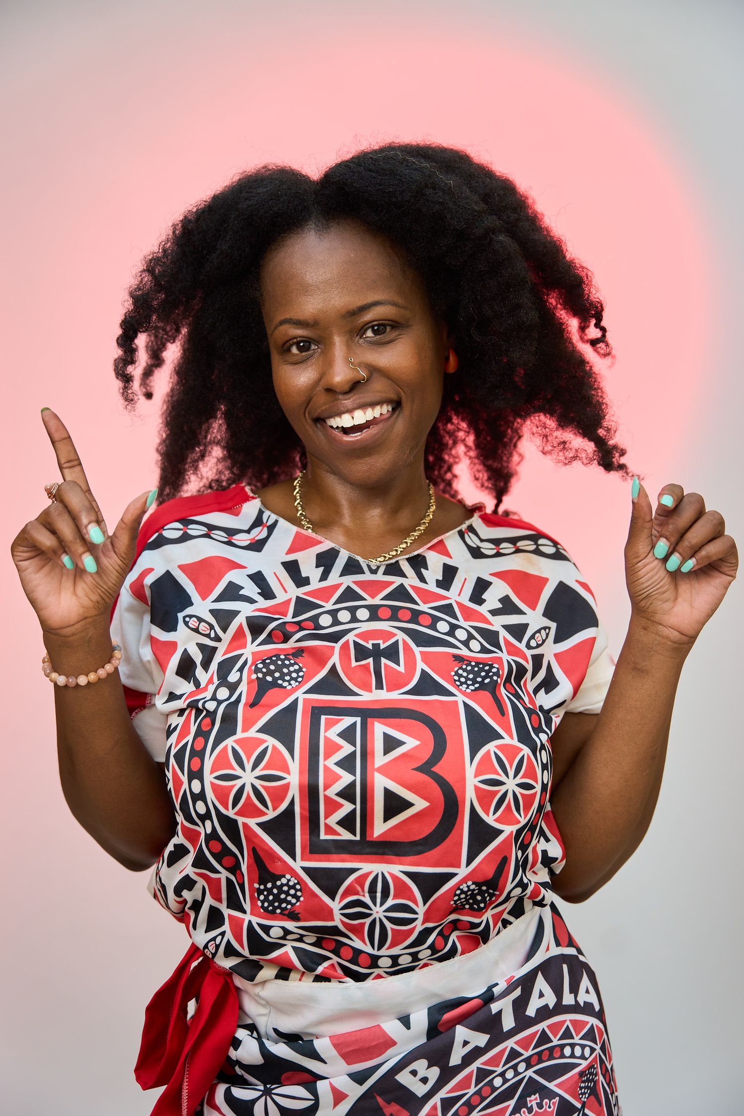 Smiling woman in colorful Batala patterned shirt pointing upward against a pink background.