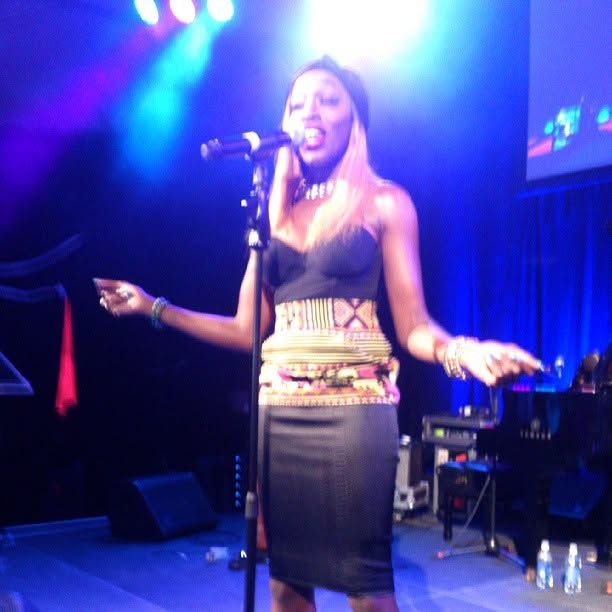 Female singer in a black top and patterned skirt performs on stage, singing into a microphone under blue and purple lights.